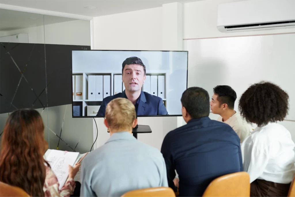 A diverse group of colleagues in a bright office sitting in front of a large screen to watch a key presentation from a GlobalMeet Webcast