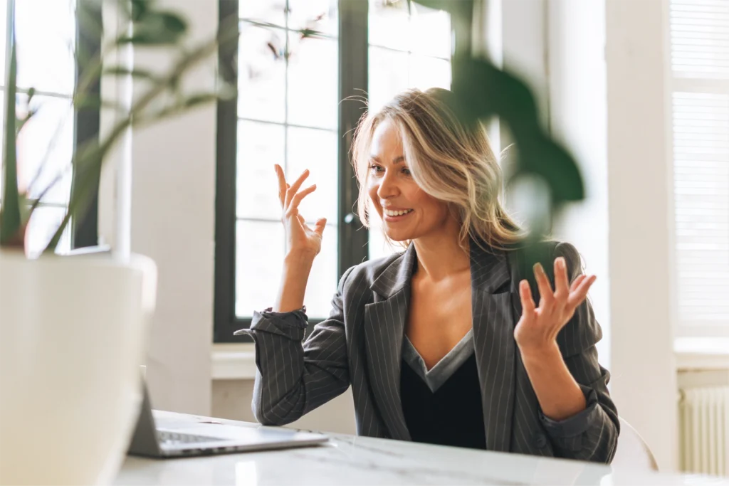 A smiling woman sitting at a bright desk engaging with a laptop