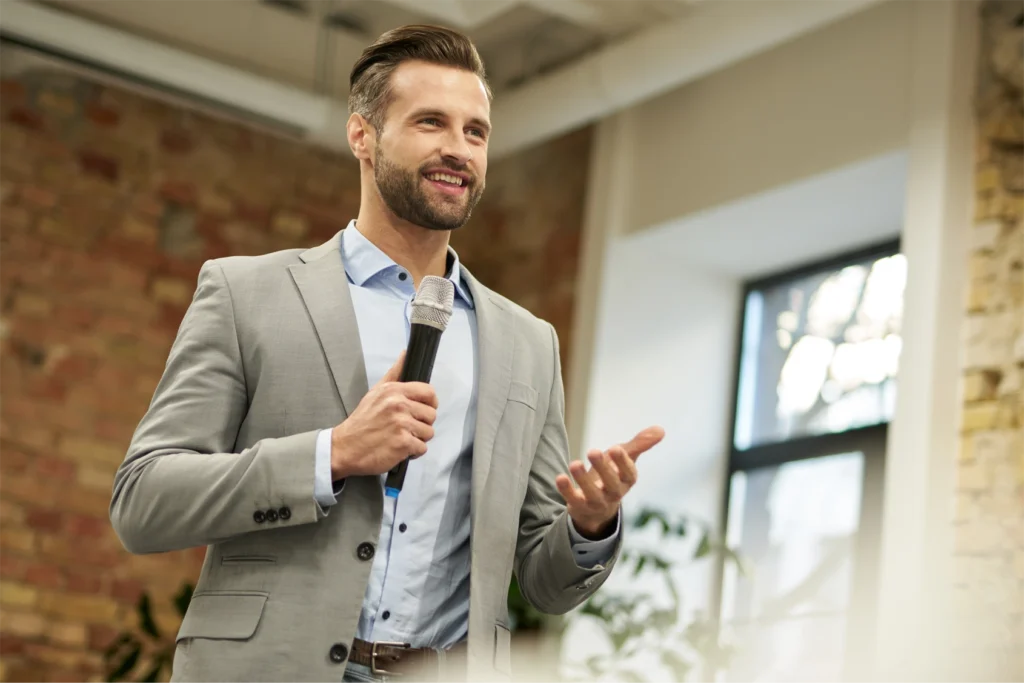 A man in a suit standing in a well lit room holding a microphone and smiling